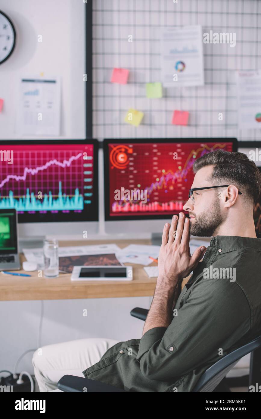 Side view of serious data analyst sitting at table with computers in office Stock Photo