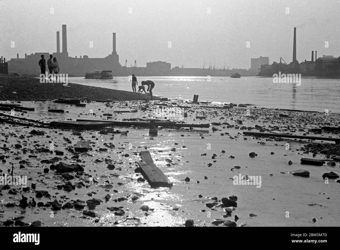 Historic London:  Mudlarking at low water beside the River Thames near Greenwich Pier on a summer’s evening in July 1969.  Passenger craft brought trippers to the pier from up river.  In the background, the coal-fired Deptford Power Station, site of the first modern high-voltage power station in the world.    At one time the second largest station in Britain, the final chimney came down in 1992 and the site is now part of the Millennium Quay development, which includes more than 600 apartments. Stock Photo
