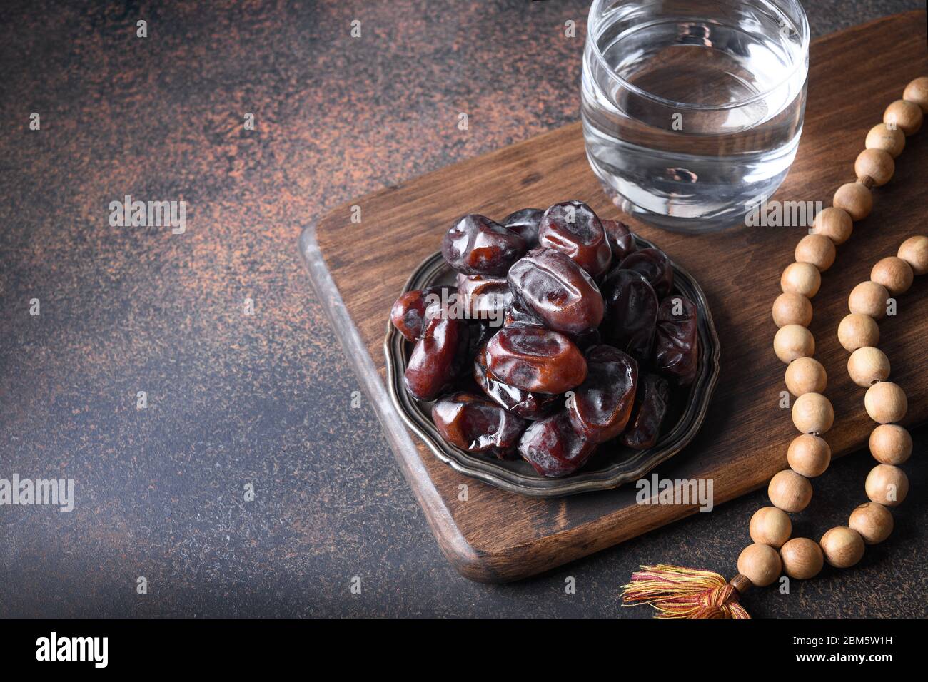 Ramadan. Dates in bowl, glass of water, wooden prayer beads on brown. Eid Mubarak. Religious tradition. Close up. Stock Photo
