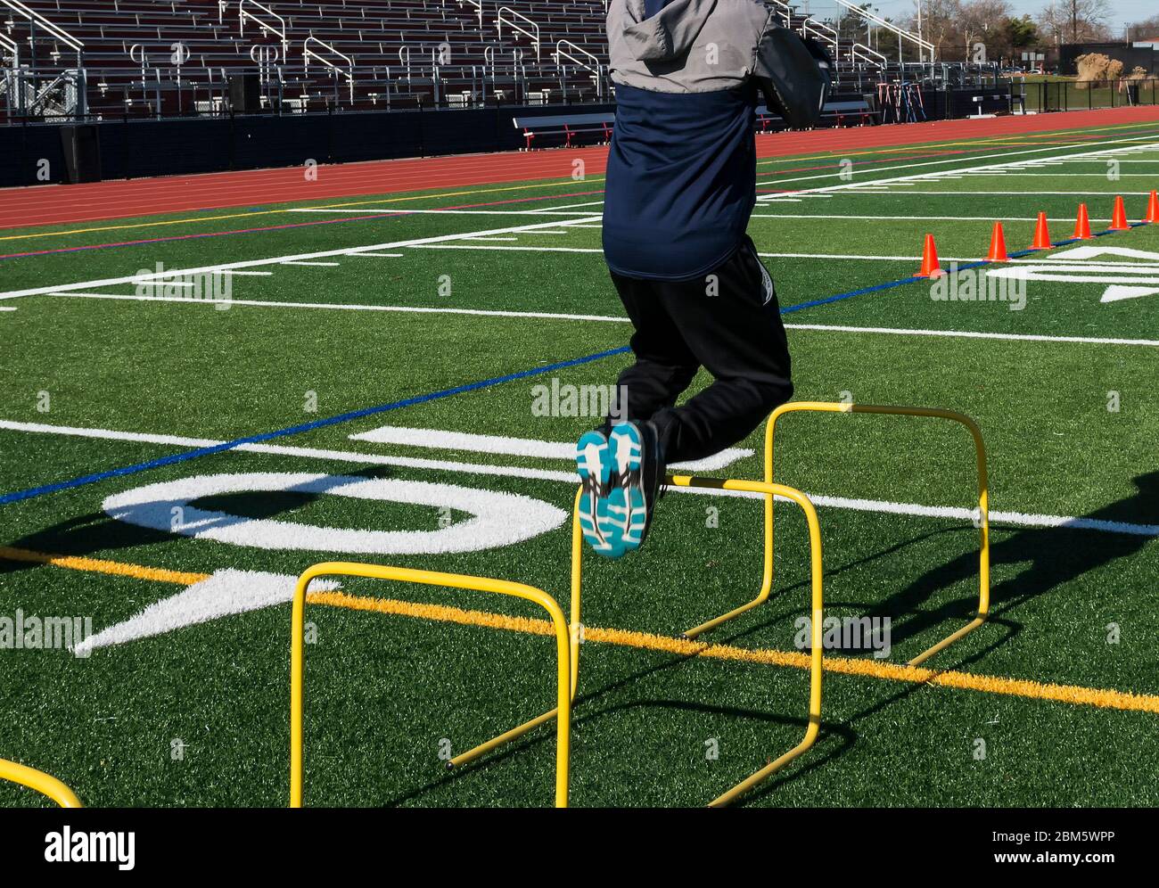 A track and field athlete jumping over yellow mini hurdles on a turf field from behind the runner. Stock Photo