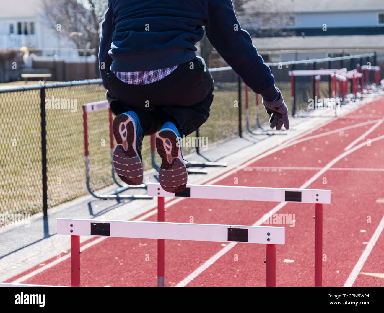 A high school male athlette is jumping over hurdles on a cold afternoon