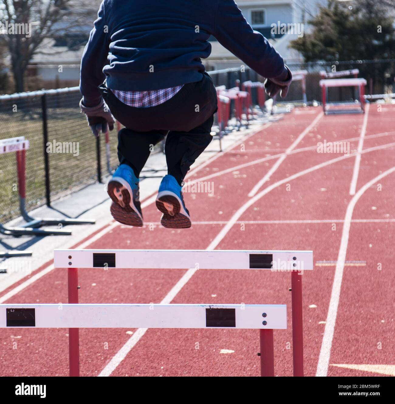 A track and field runner is jumping over hurdles on a red track during winter with gloves on. Stock Photo