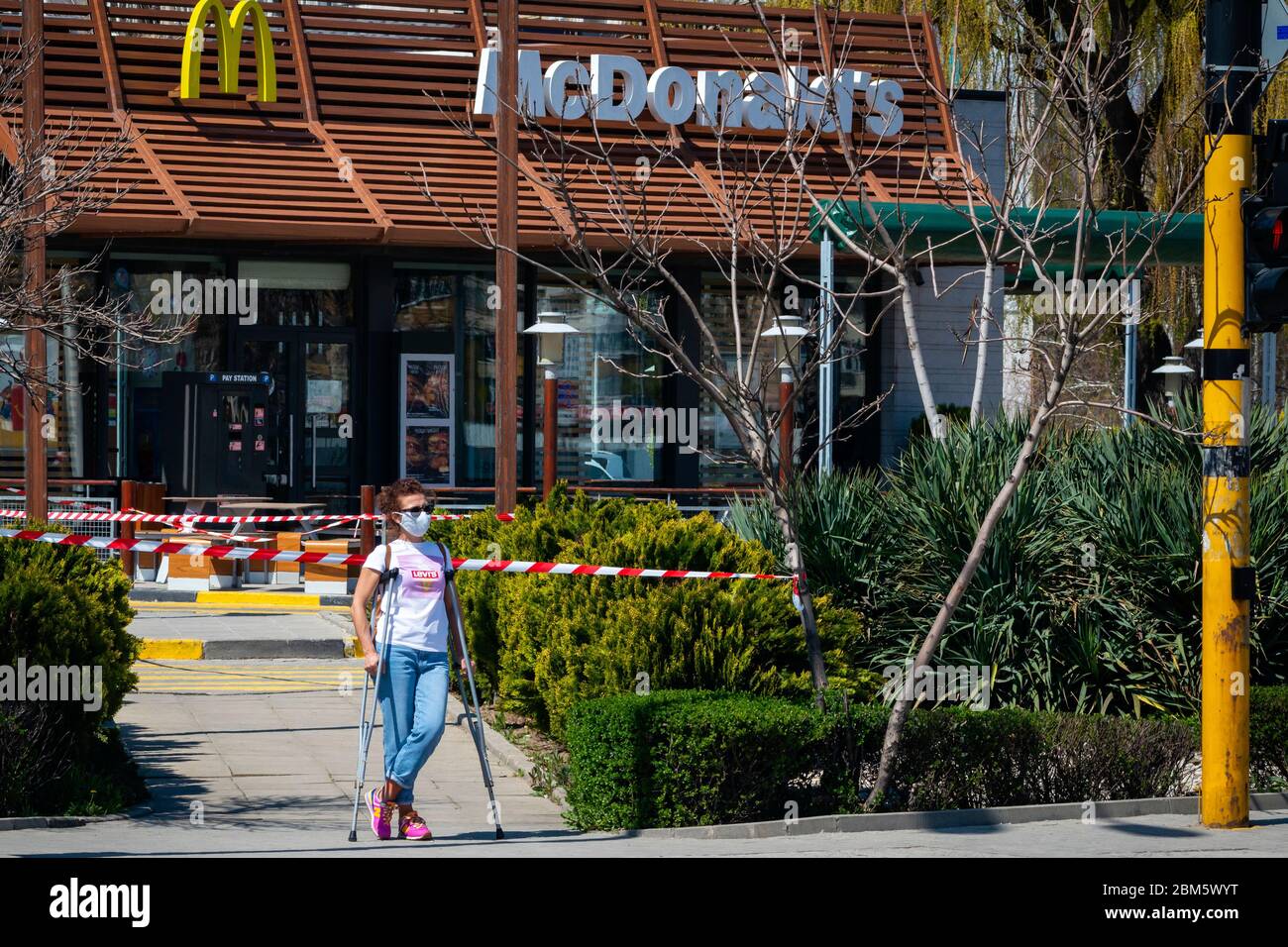 Woman with crutches wearing protective face mask standing in front of closed for normal business McDonald's fast food restaurant allowing only drive-in or drive-thru takeaway food due to the spread of the Coronavirus Pandemic of Covid-19 in Sofia, Bulgaria as of April 2020 Stock Photo