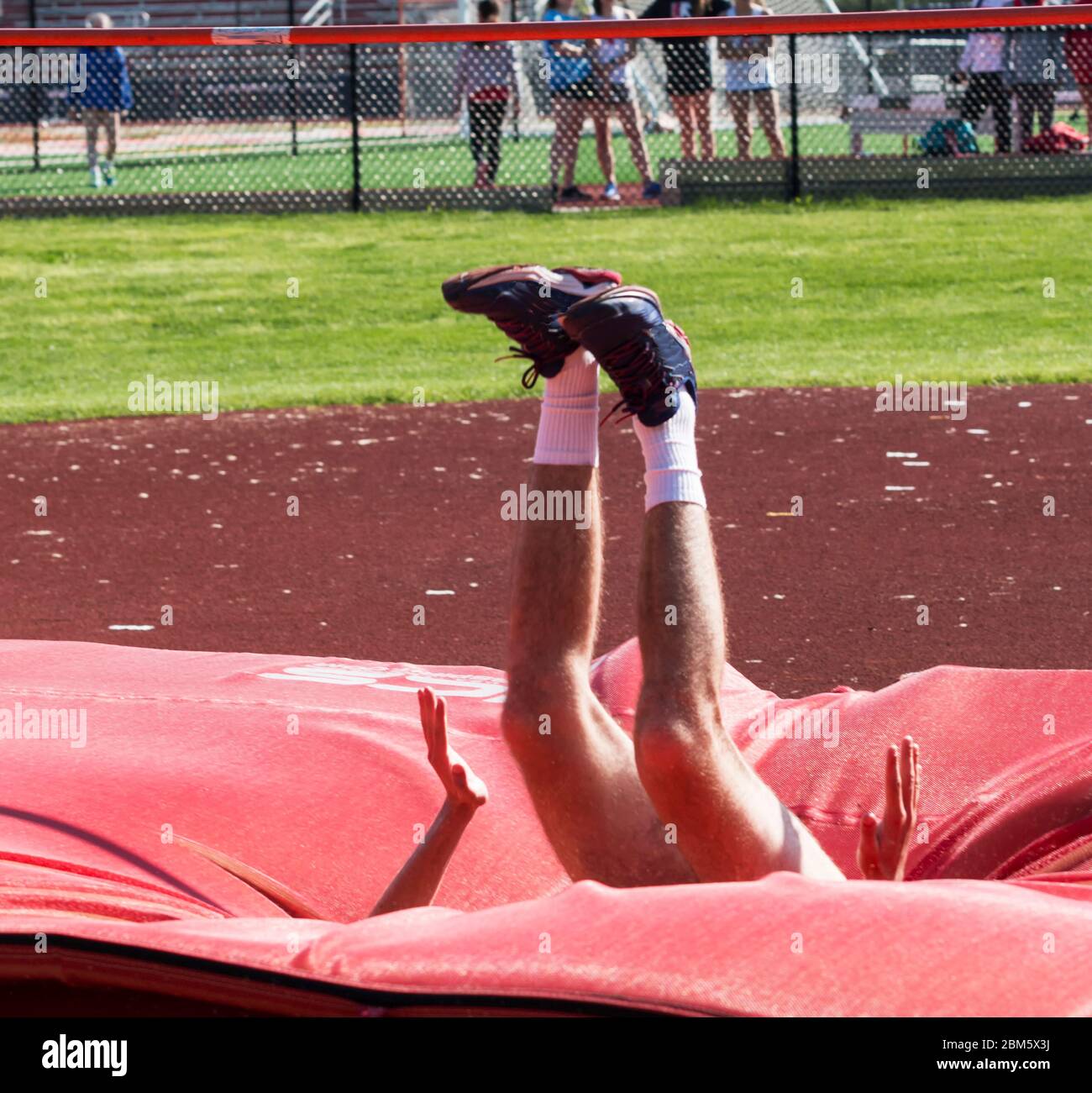 A male high school track and field athlete is engulfed by red matts as he lands on them during a high jump competition. Stock Photo
