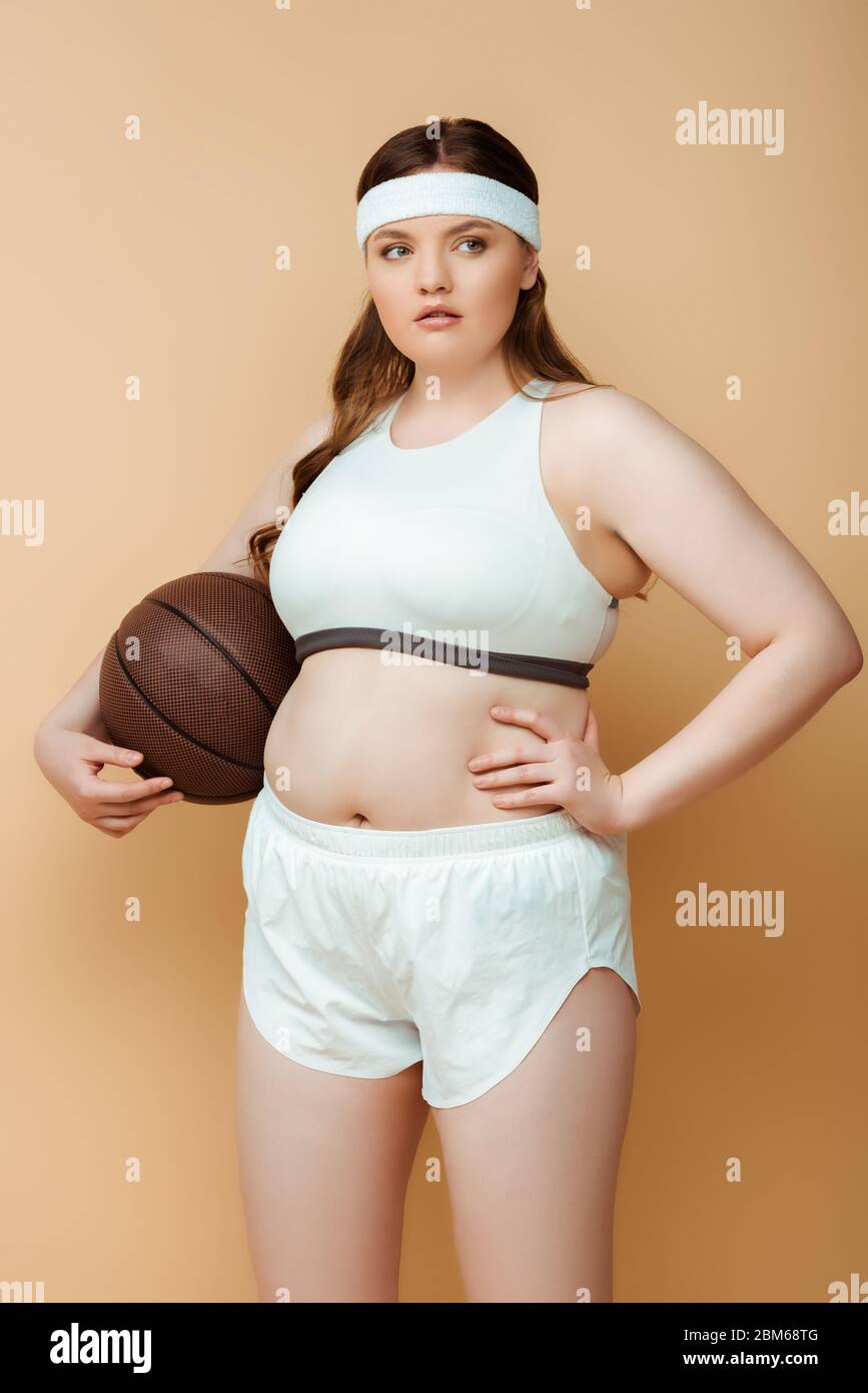 Plus size sportswoman with hand on hip looking away and holding ball on beige background Stock Photo
