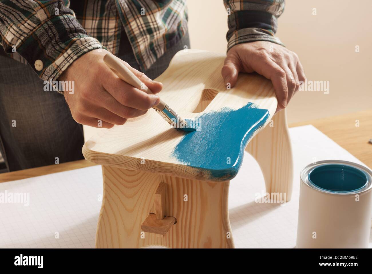 Craftsman varnishing a wooden handmade stool at home with a blue coating on a work table, hands close up Stock Photo