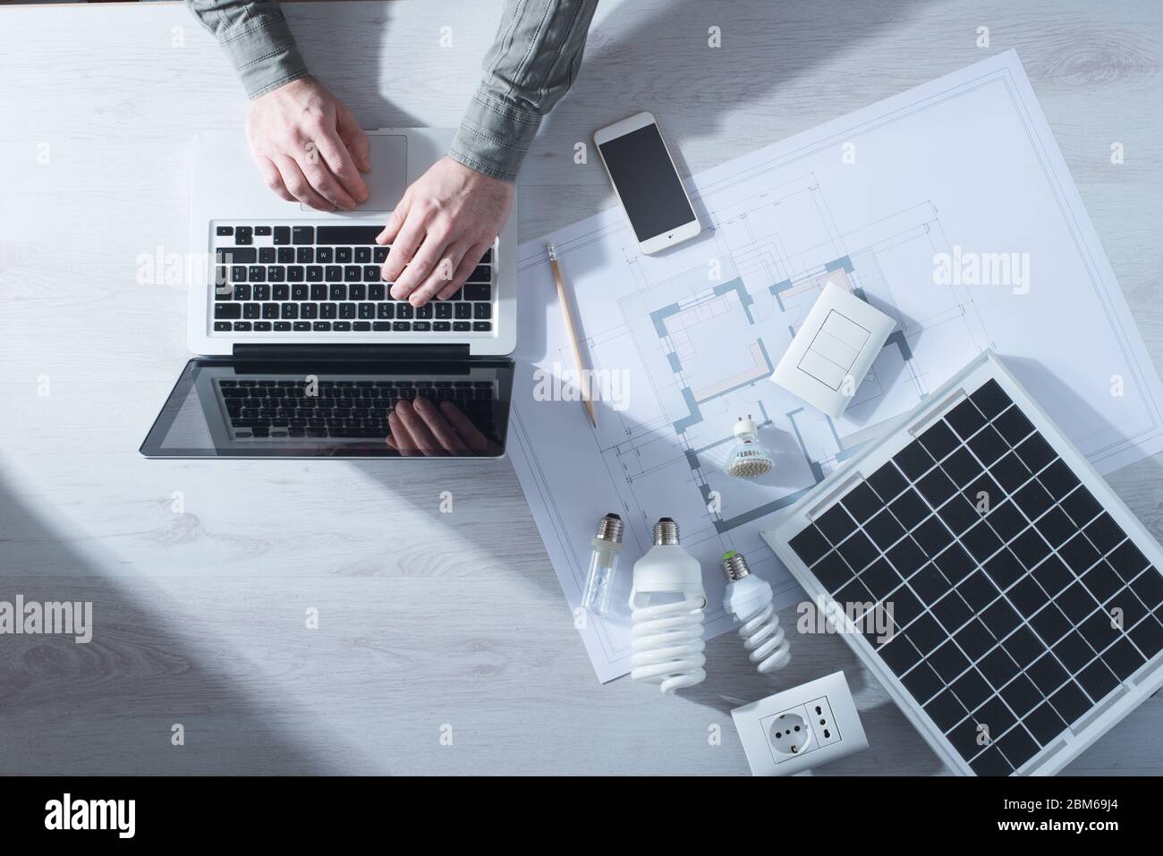 Man working at his desk on a laptop with energy saving CFL lamps, a solar panel and a house project, top view Stock Photo