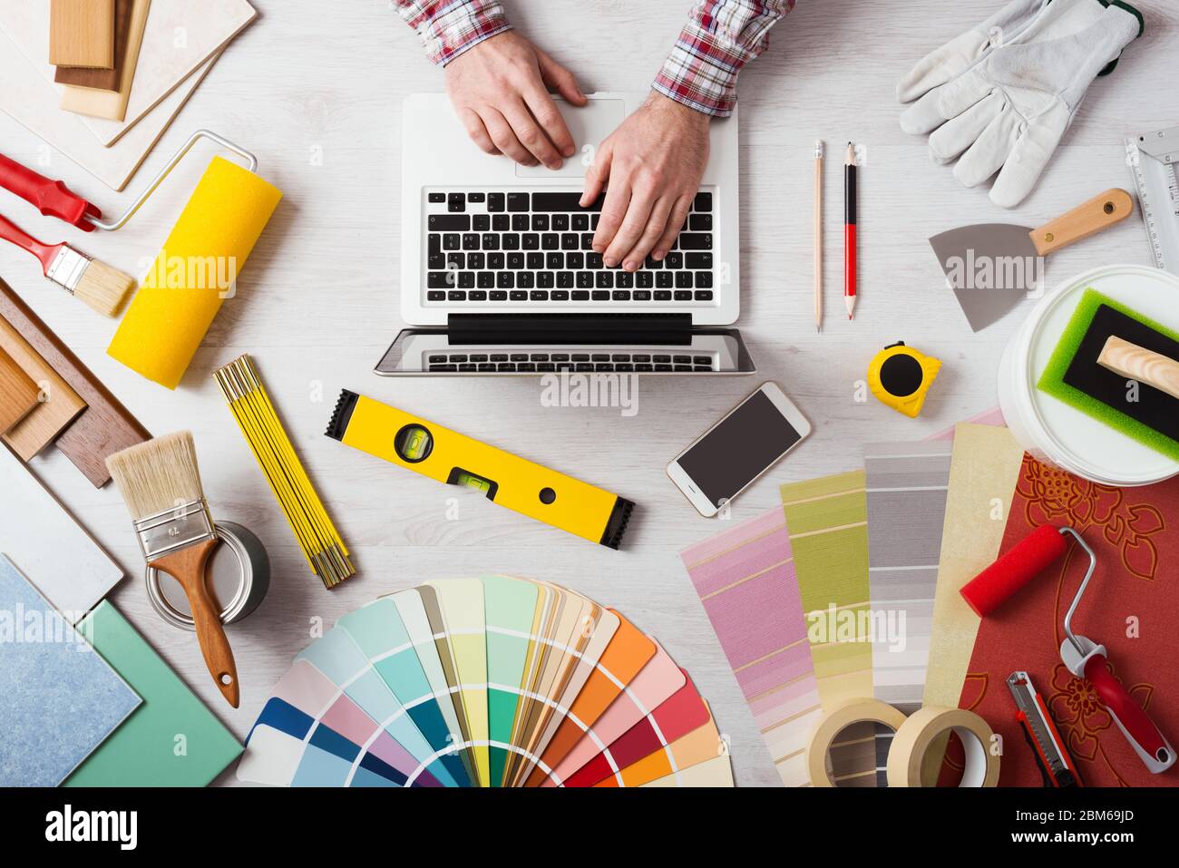 Professional decorator's hands working at his desk and typing on a laptop, color swatches, paint rollers and tools on work table, top view Stock Photo