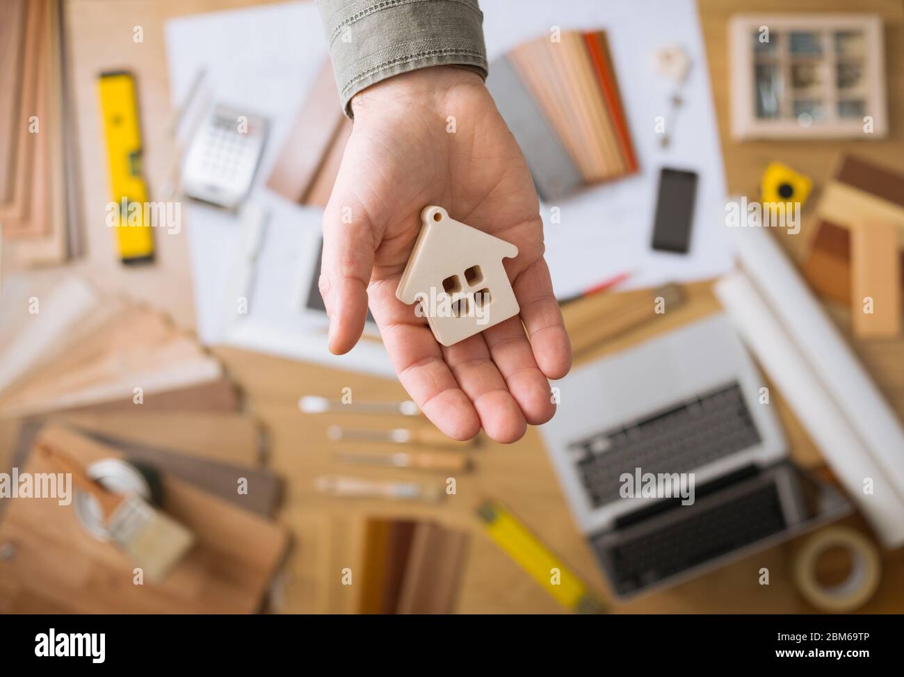 Real estate agent holding a small house, desktop with tools, wood swatches and computer on background, top view Stock Photo