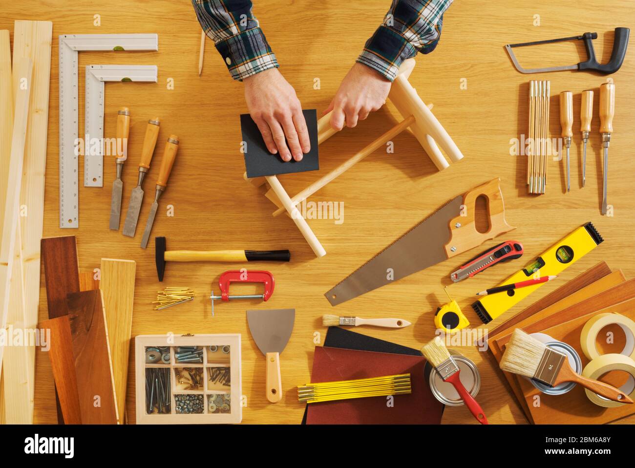 Male hands working on a footstool on a worktable smoothing the surface, DIY tools all around, top view Stock Photo