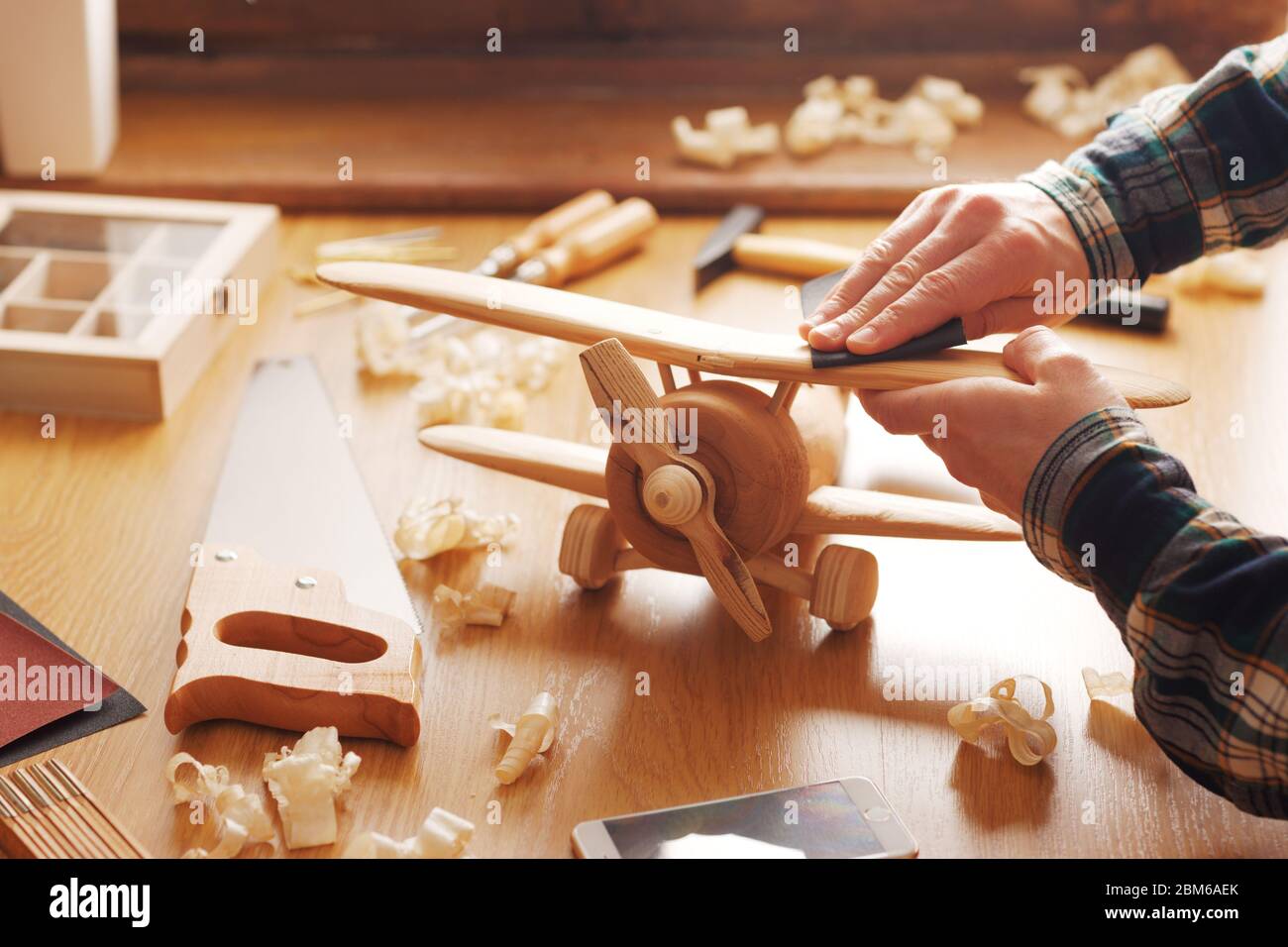 Craftsman smoothing a wooden toy surface with sandpaper, tools and wood shavings all around, hands close up Stock Photo