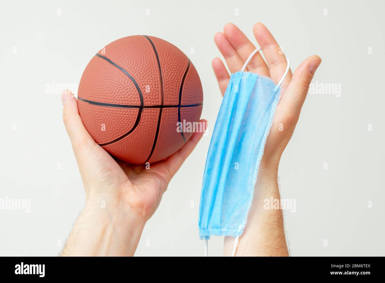 Man hands holding basketball ball with medical protective mask on light background during coronavirus. Basketball paused because coronavirus. Stock Photo