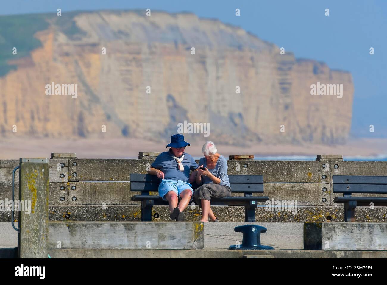 West Bay, Dorset, UK.  7th May 2020. UK Weather.  A couple sitting on a bench on the pier at the seaside resort of West Bay in Dorset enjoying the hot sunshine during the coronavirus pandemic lockdown.   Picture Credit: Graham Hunt/Alamy Live News Stock Photo