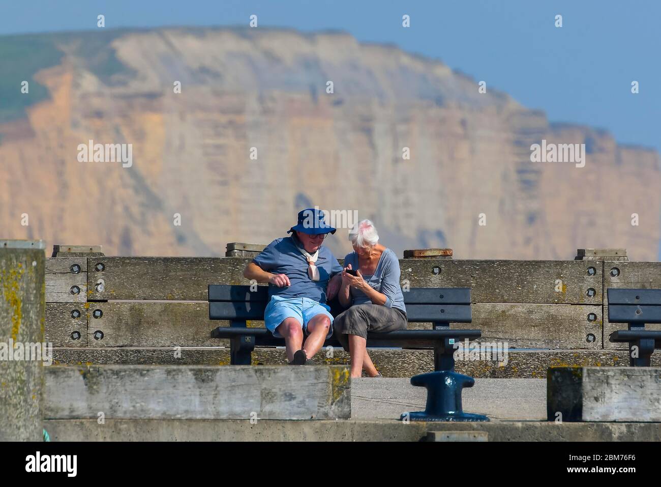 West Bay, Dorset, UK.  7th May 2020. UK Weather.  A couple sitting on a bench on the pier at the seaside resort of West Bay in Dorset enjoying the hot sunshine during the coronavirus pandemic lockdown.   Picture Credit: Graham Hunt/Alamy Live News Stock Photo
