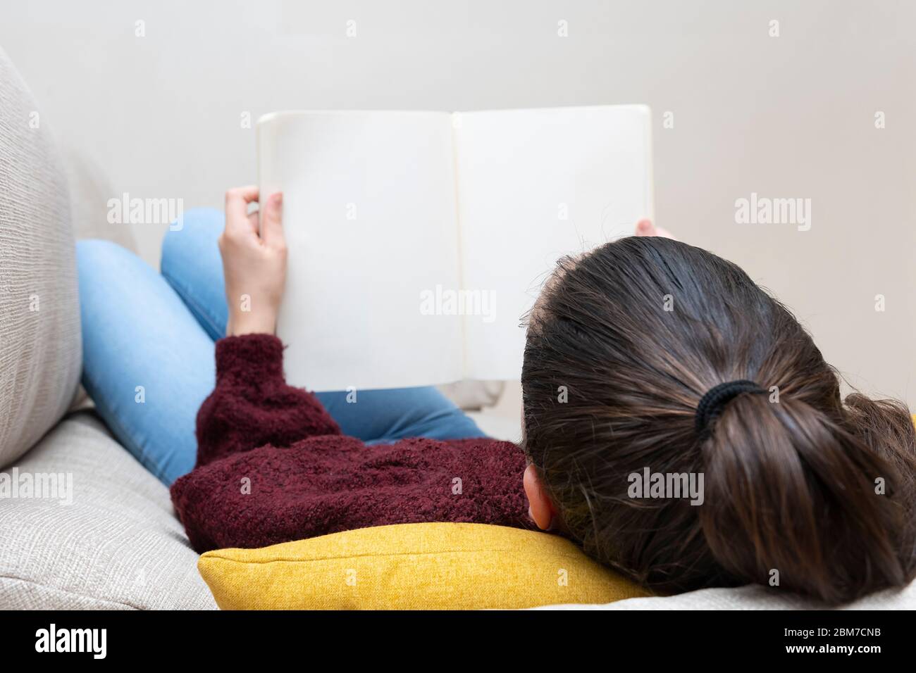 Girl with tied hair lying on a sofa with her head on a cushion reading a book Stock Photo