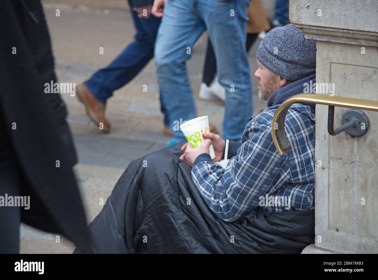 A homeless man sitting in a sleeping bag and holding a cup for