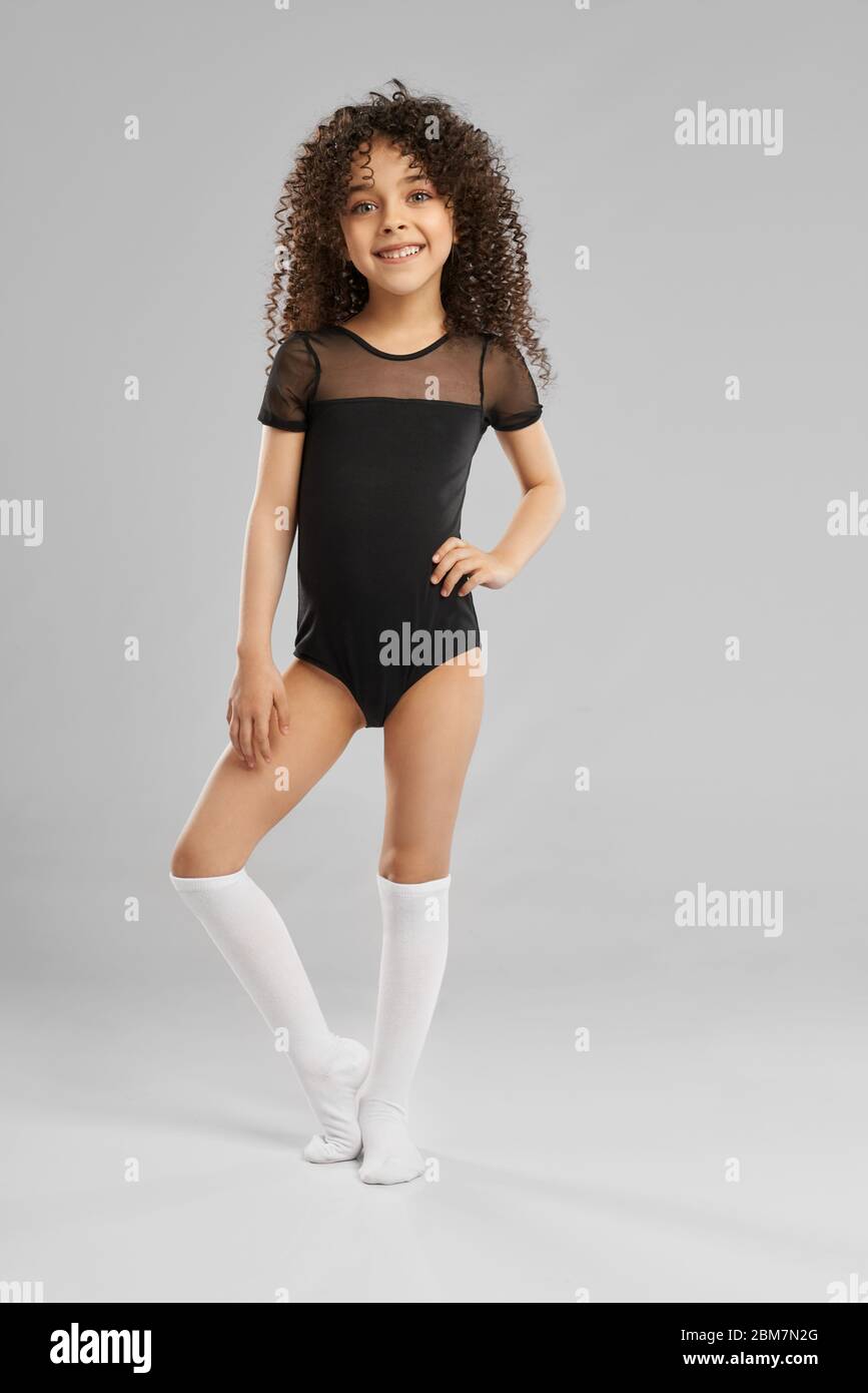 Full length portrait of adorable smiling girl in leotard and white knee socks posing with hand on waist, isolated on gray studio background. Little female model with curly hair looking at camera. Stock Photo