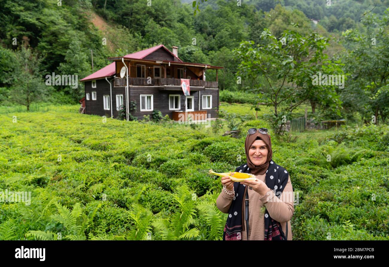 Rize / Turkey - August 06 2019: Tourist Turkish woman eats boiled corn and wooden house with tea field just near  Senyuva (Cinciva) bridge over Firtin Stock Photo