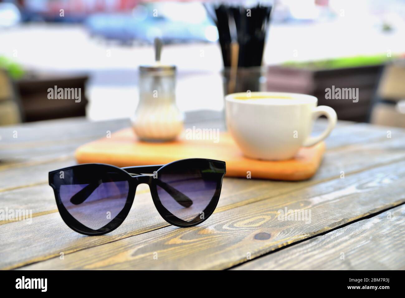 cup of coffee on a wooden stand is on the table in a cafe summer morning, sunglasses in the foreground, close-up Stock Photo