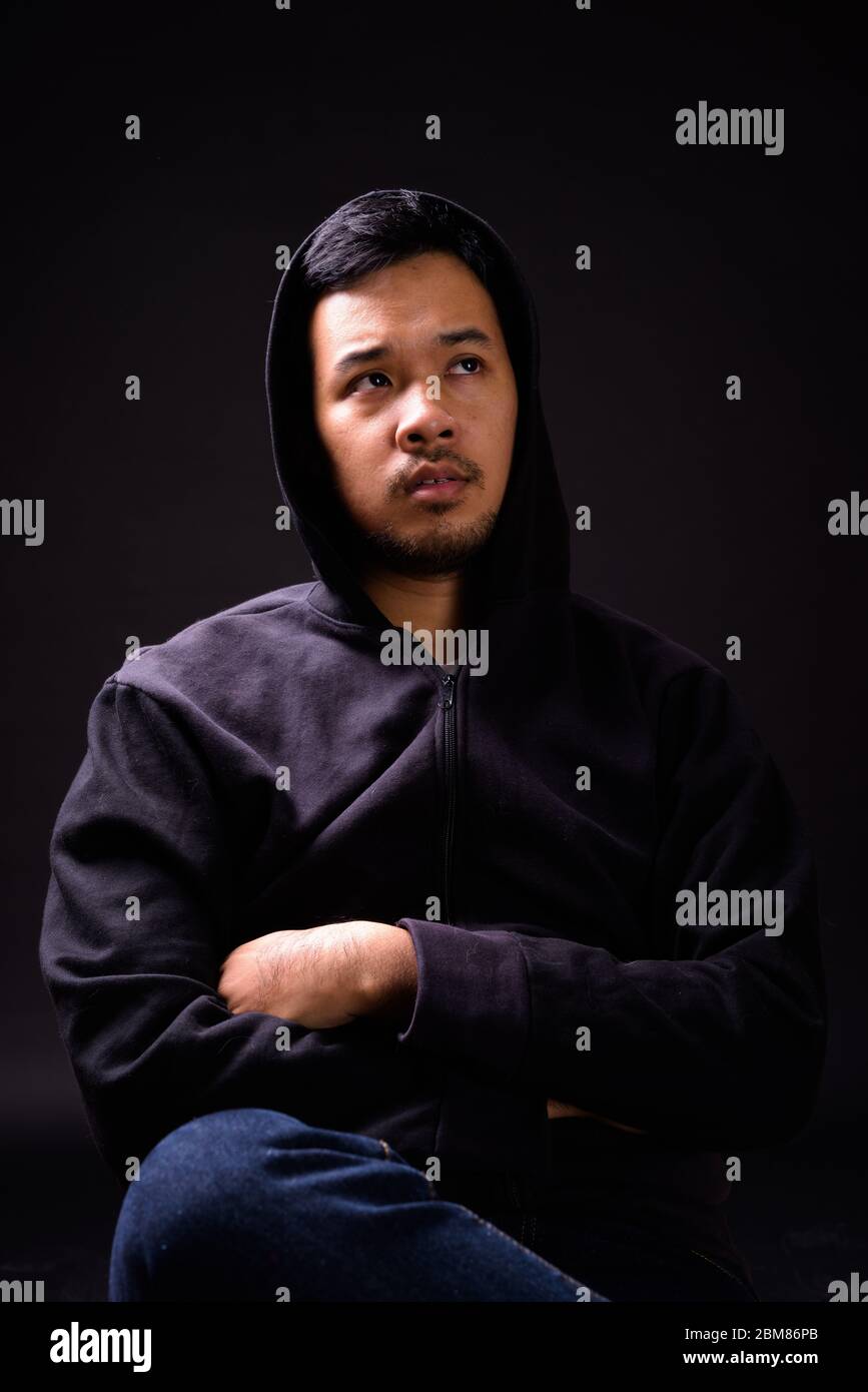 Portrait of young Asian man with hoodie thinking while sitting on chair ...