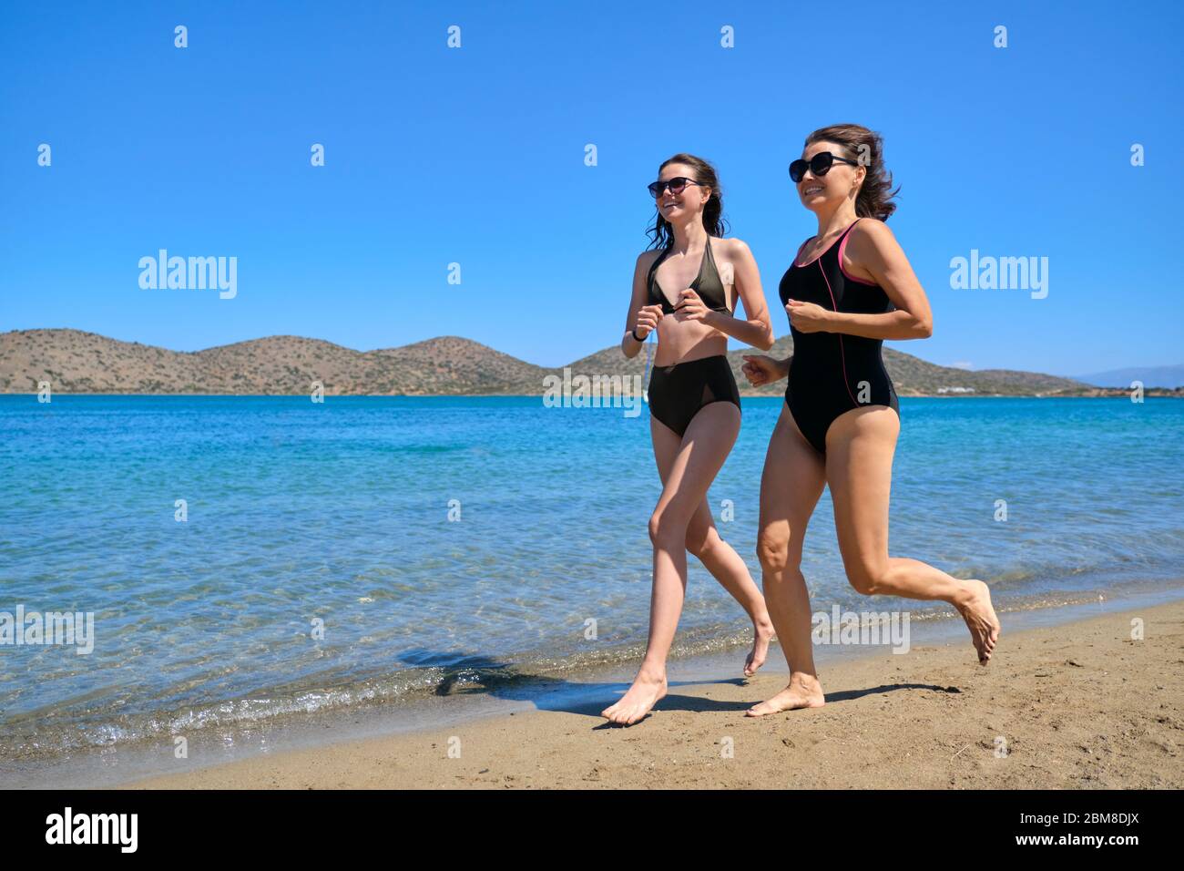 Mother and teen daughter together on beach, running by sea in swimsuit Stoc...