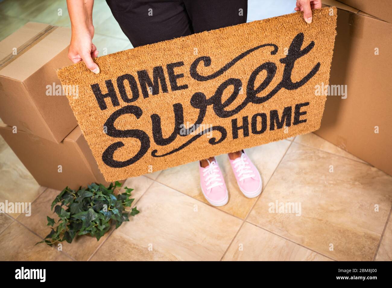 Woman in Pink Shoes Holding Home Sweet Home Welcome Mat, Boxes and Plant. Stock Photo