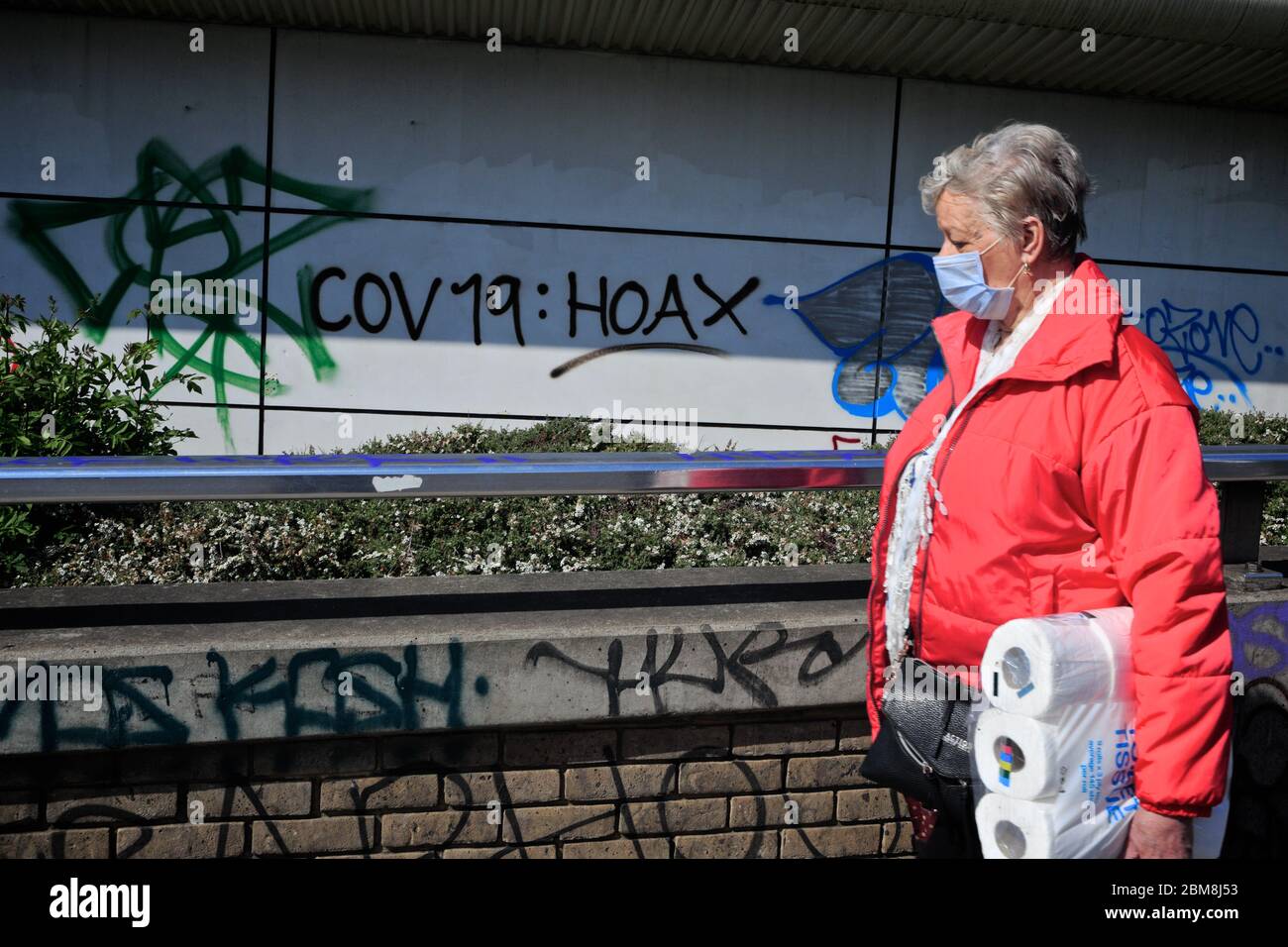 London, UK.  A woman carrying toilet roll, and wearing face covering, can be seen walking past 'covid- 19 hoax' graffiti amid the covid world crisis. Stock Photo