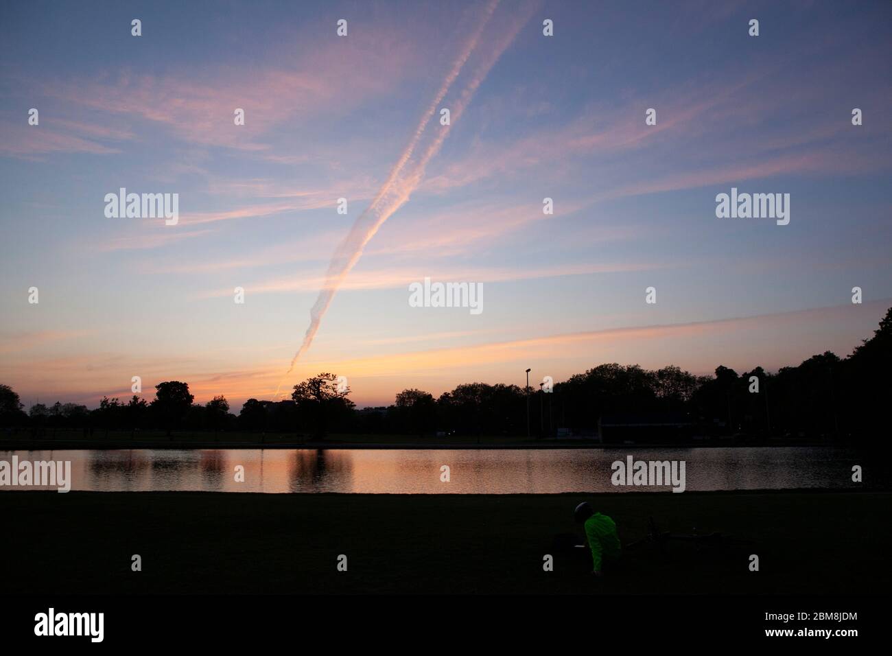 London, UK, 7 May 2020: After days of clear skies increasing number of flights are leaving vapour trails in the sky. On Clapham Common the resulting dramatic sunset was reflected in the Lond Pond. Anna Watson/Alamy Live News Stock Photo