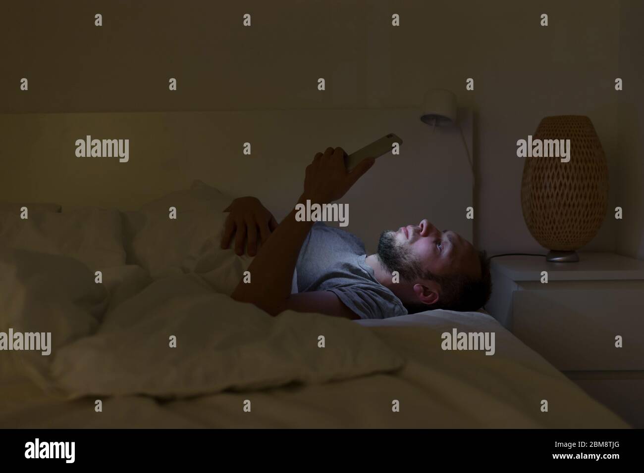 Mobile addiction, nomophobia, insomnia, sleep disorder concept. Bored man chatting and surfing on internet with smart phone late at night, lying on be Stock Photo