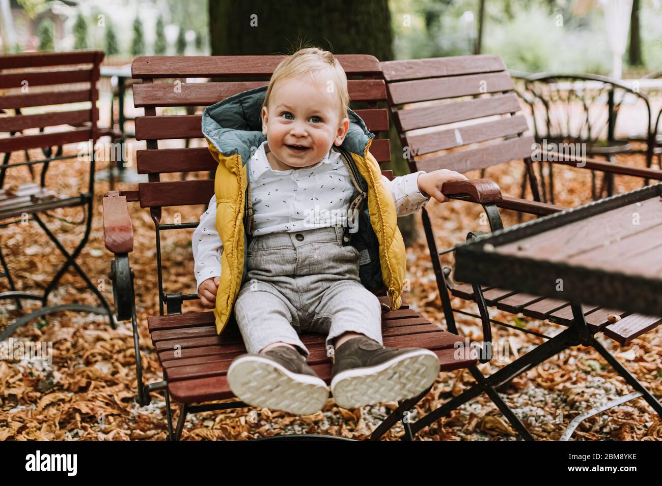 1 year old cute boy in stylish yellow jacket smiling and sitting on the chair in the park Stock Photo