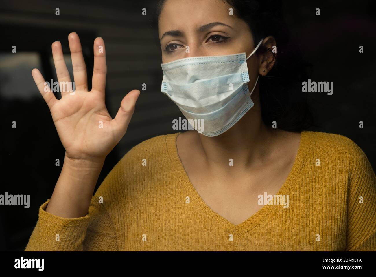 Young sad woman wearing a face mask looking through the window, self isolated at home. Coronavirus and Quarentine concept Stock Photo
