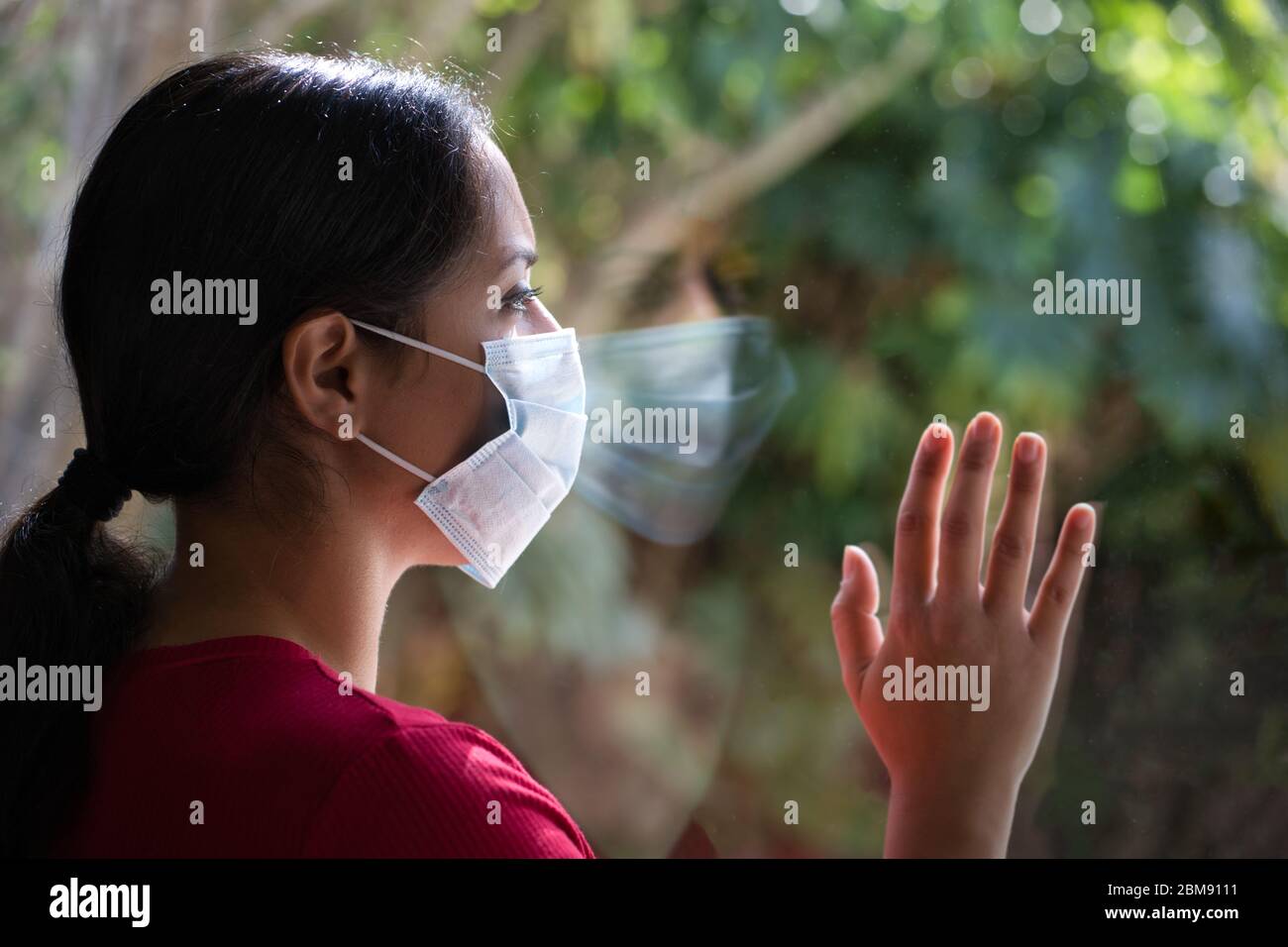 Brunette woman wearing a face mask looking through the window with her reflection on the glass, self isolated at home. Coronavirus and Quarentine conc Stock Photo