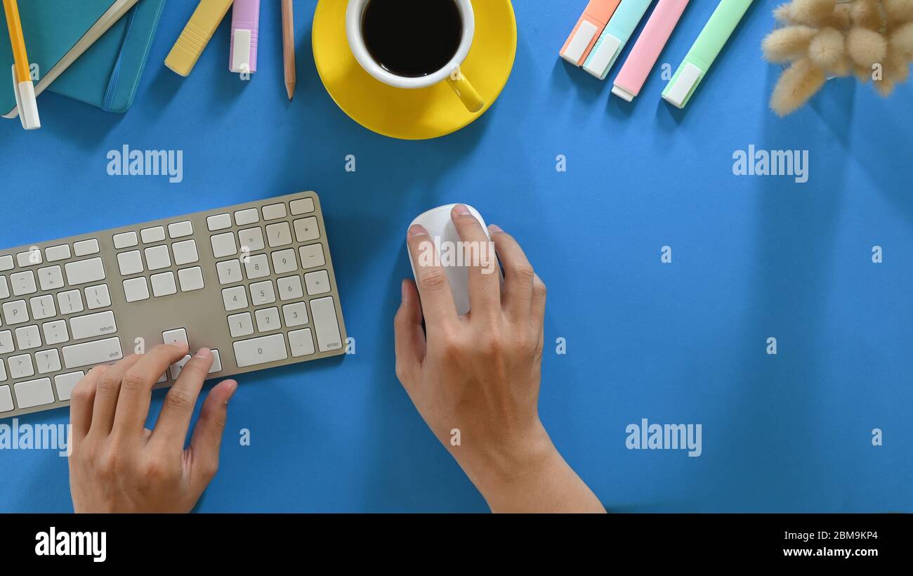 Top view image of hands while using a wireless keyboard and mouse that putting on colorful working desk and surrounded by hot coffee cup, marker pens, Stock Photo