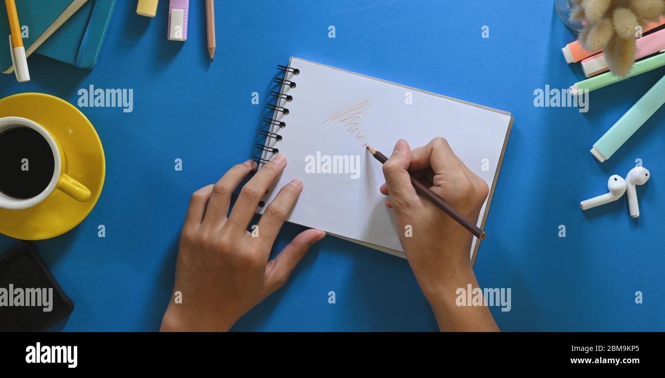 Top view image of hands while taking note on notebook that putting on colorful working desk and surrounded by hot coffee cup, marker pens, wireless ea Stock Photo