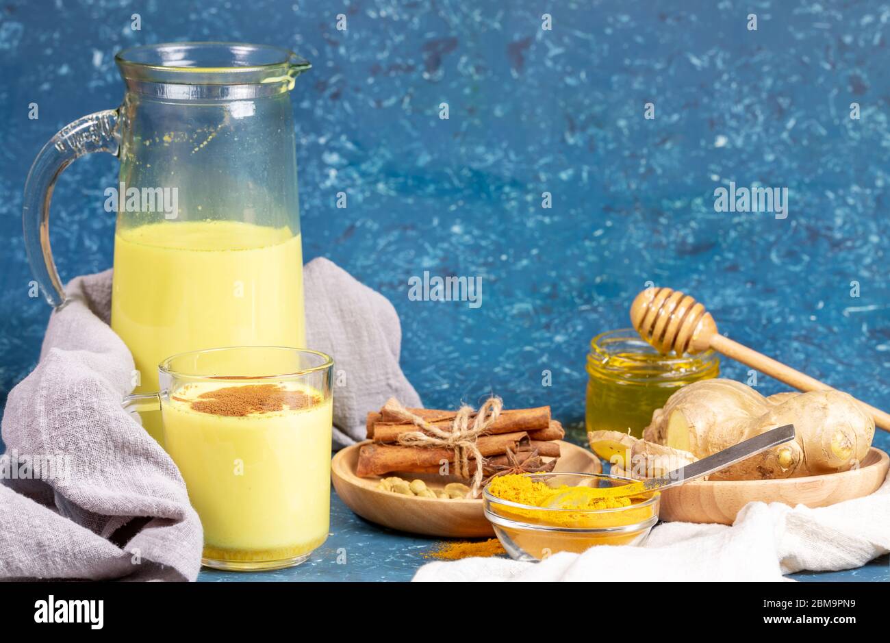 Organic turmeric milk. Composition of glass and decanter with golden milk, ingredients and honey on blue background. Concept of healthy eating, diet a Stock Photo