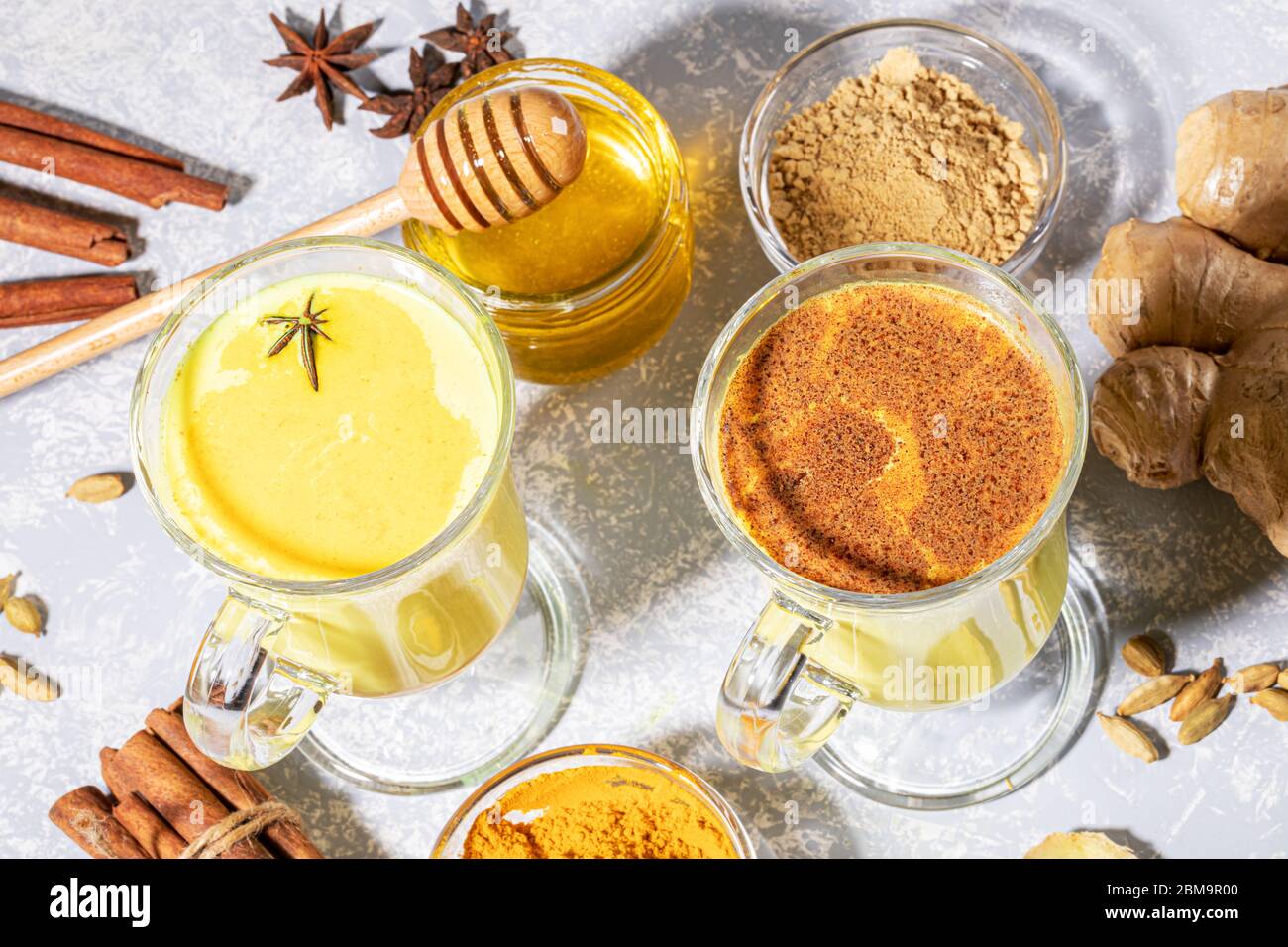 Organic turmeric milk. Two glasses for mulled wine with golden milk with ingredients on light grey backdrop with shadows. Alternative medicine. Health Stock Photo