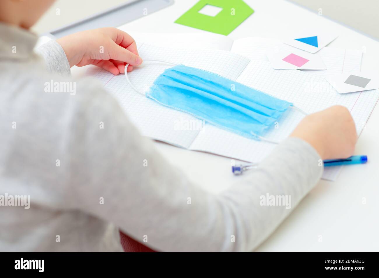 Hands of child holding a medical mask over copybook during studying at home. Home learning concept. Stock Photo