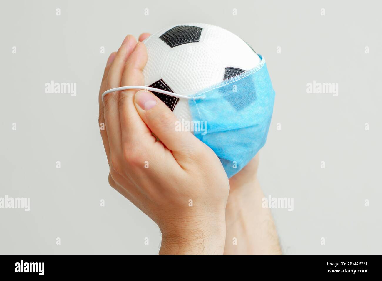 Man's hands holding soccer ball with a medical blue mask on a light background with copy space. Cancellation of football competitions. Stock Photo