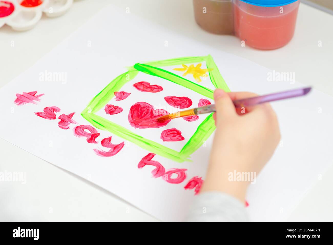 Child is drawing red hearts inside house on white sheet of paper with words Stay Home. Stay Home concept. Children creativity. Stock Photo