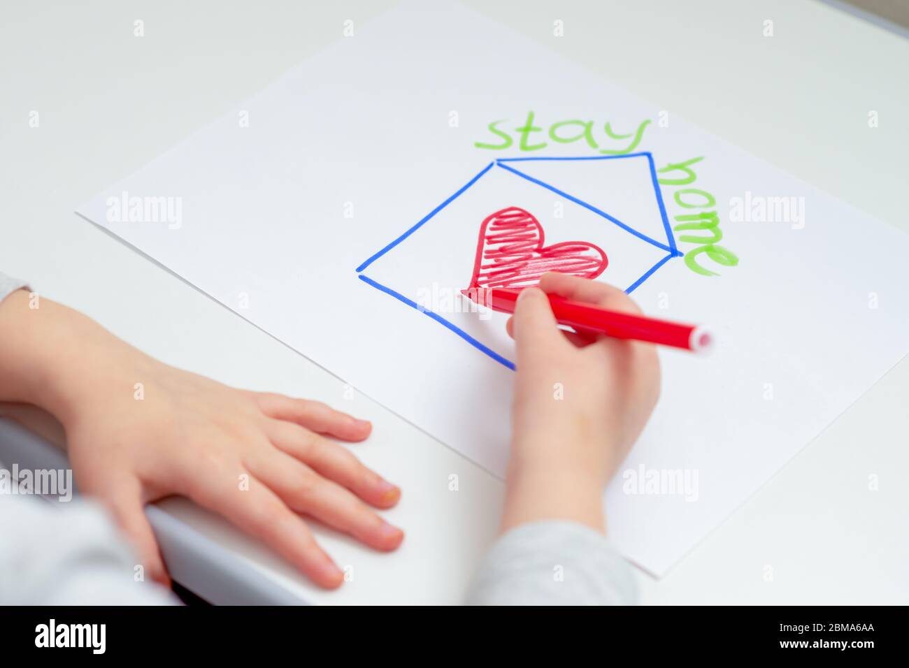 Hands of child drawing red heart inside house with words Stay Home on white sheet of paper. Stay Home concept. Stock Photo