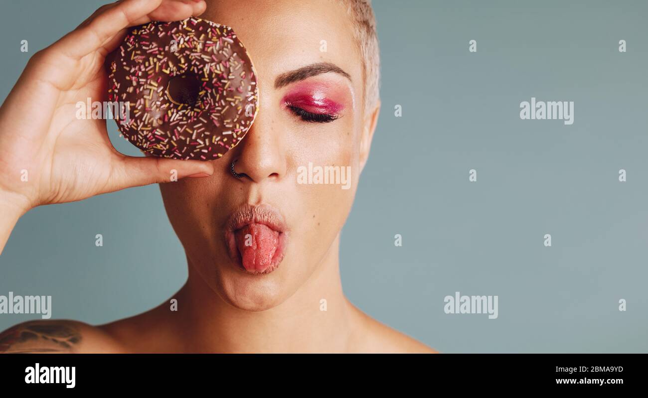 Close-up of a woman with short hair holding a donut and sticking out tongue. Female model a donut in front of her face on grey background. Stock Photo