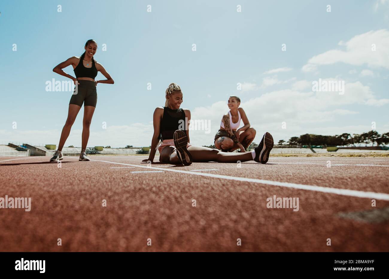 Female athletes taking a break after workout sitting on the running track in the stadium. Women relaxing and smiling after training. Stock Photo