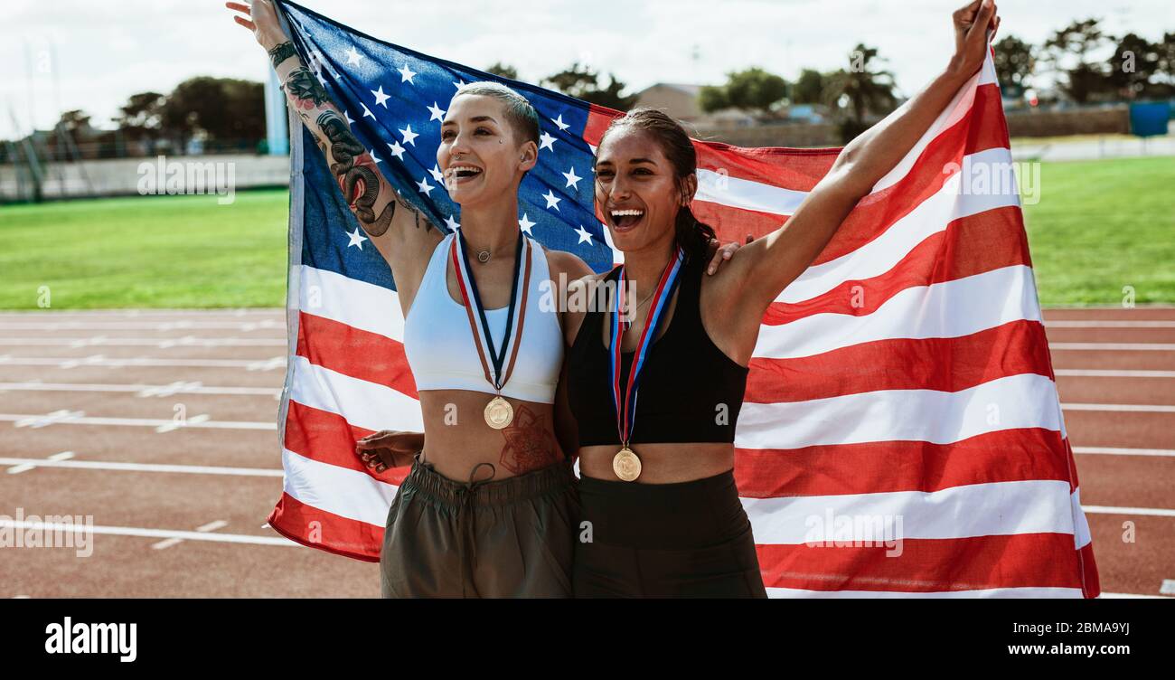 Female athletes on running track celebrating victory holding american flag. Sprinter carrying US flag and screaming after winning the sports event. Stock Photo