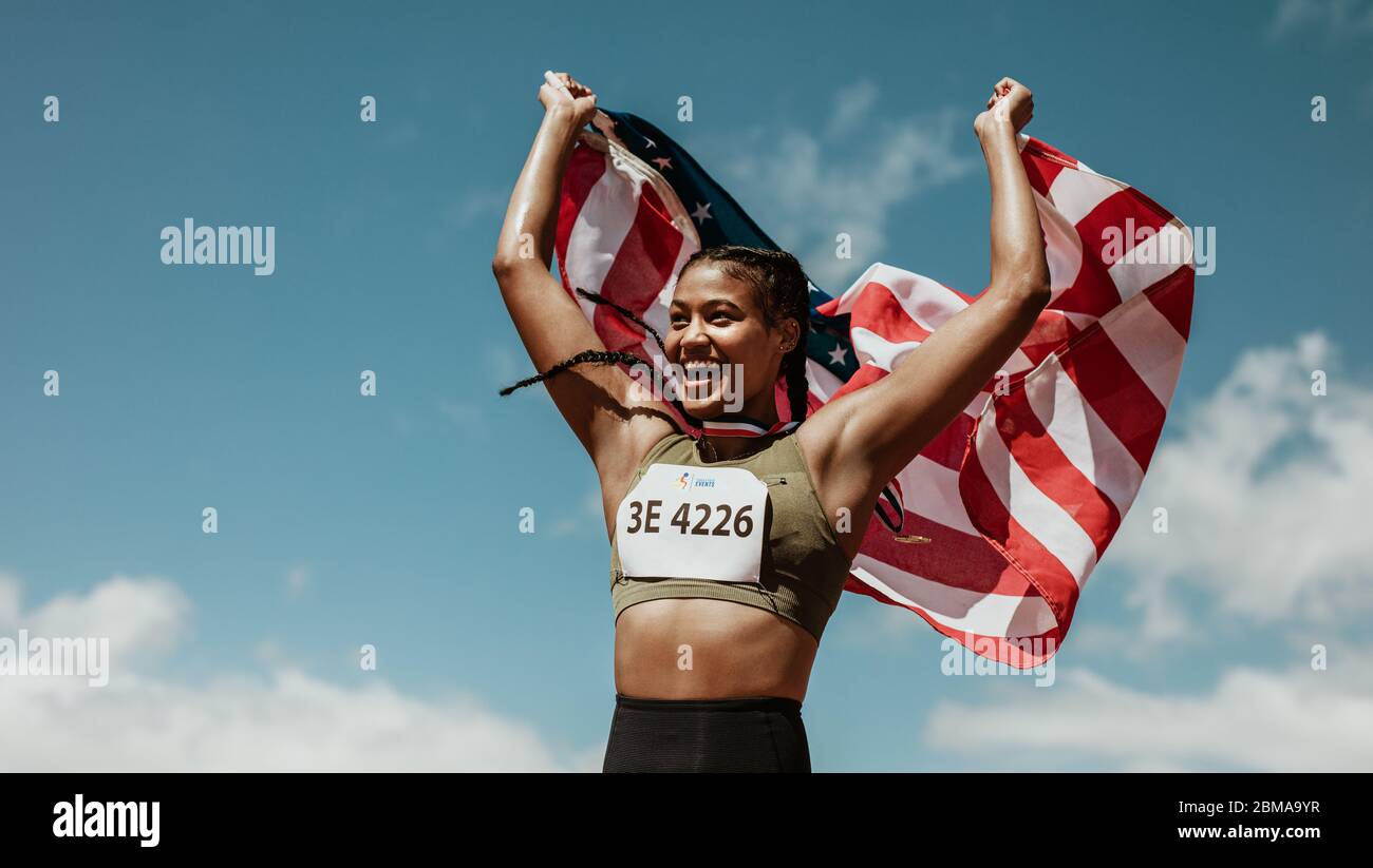 Athlete holding the american flag over the head and smiling. Female runner celebrating victory outdoors holding the US flag. Stock Photo