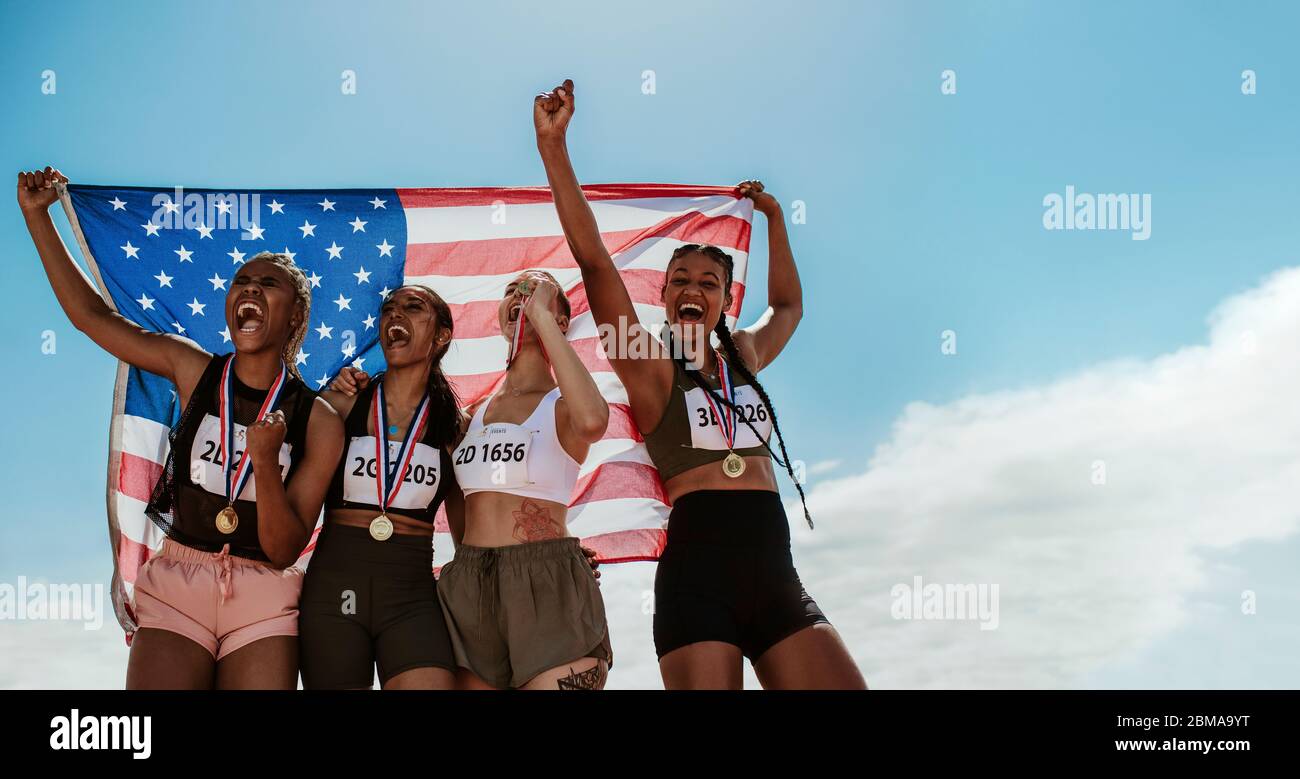 Happy multiracial athletes celebrating victory while standing together outdoors holding American flag. Group of USA runners with medals winning a comp Stock Photo