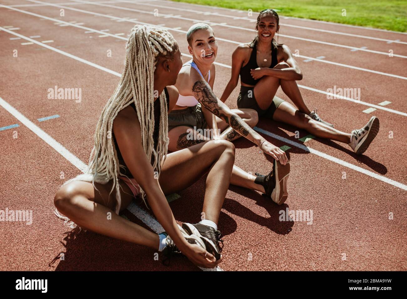 Happy group of female athletes sitting on a running track and stretching legs. Women runners doing warmup exercises at the stadium. Stock Photo