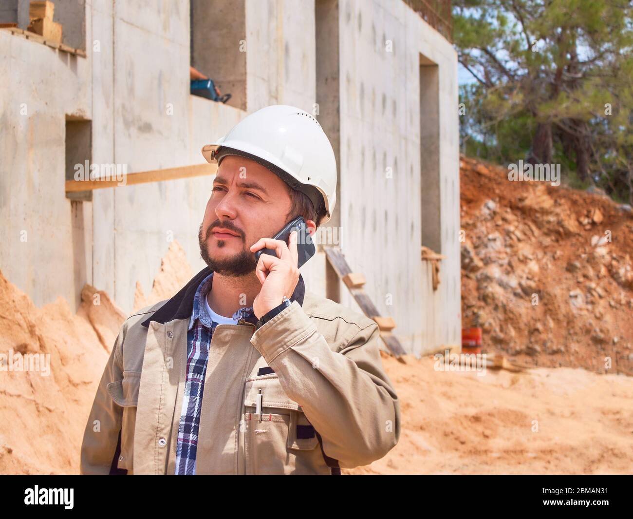 Caucasian bearded civil engineer or construction worker in white hardhat makes phone calls opposite construction site Stock Photo