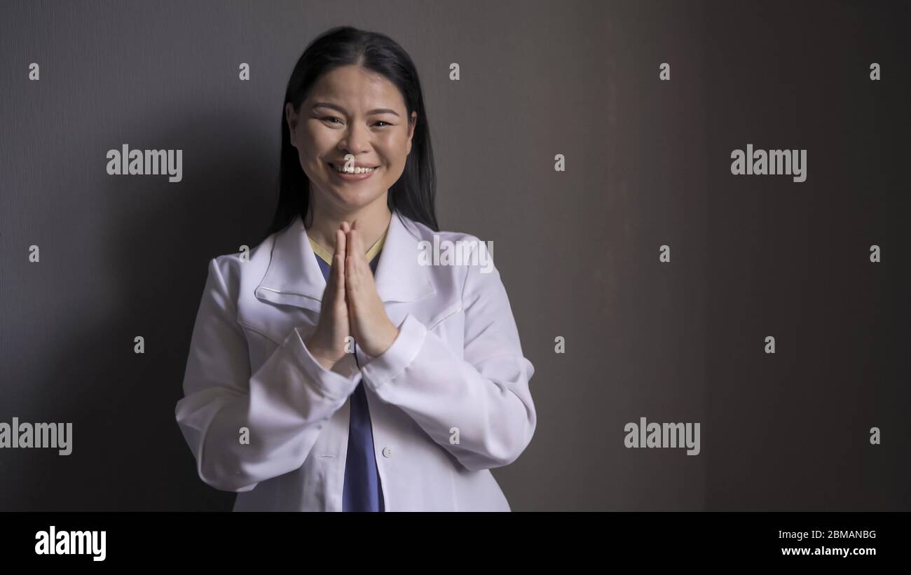 Happy doctor claps her hands toothy smiling standing on gray background. Beautiful Female medic took off the mask rejoicing at good news about the end Stock Photo