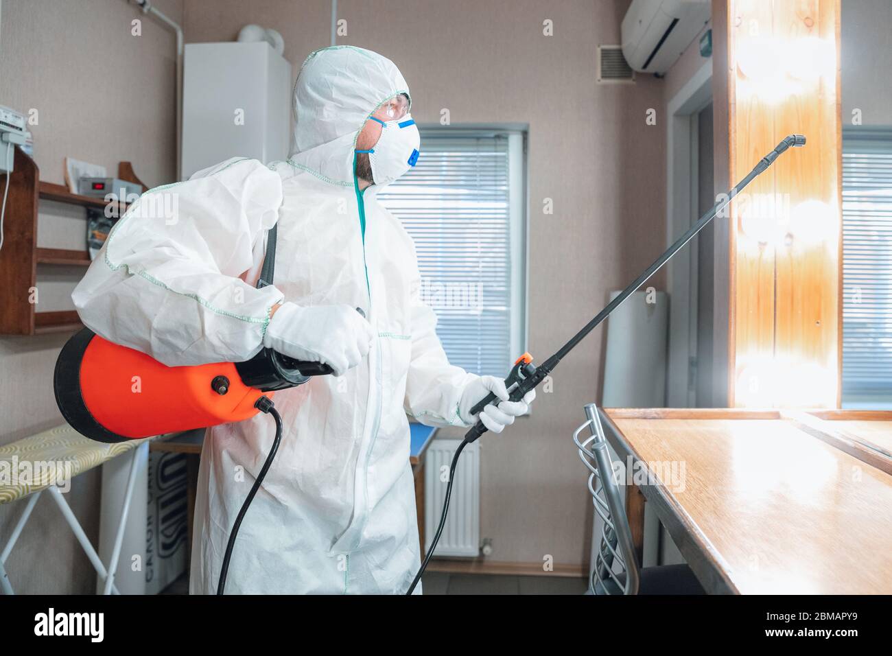 Coronavirus Pandemic. A disinfector in a protective suit and mask sprays disinfectants in house or office. Protection against COVID-19 disease. Prevention of spreding pneumonia virus with surfaces. Stock Photo