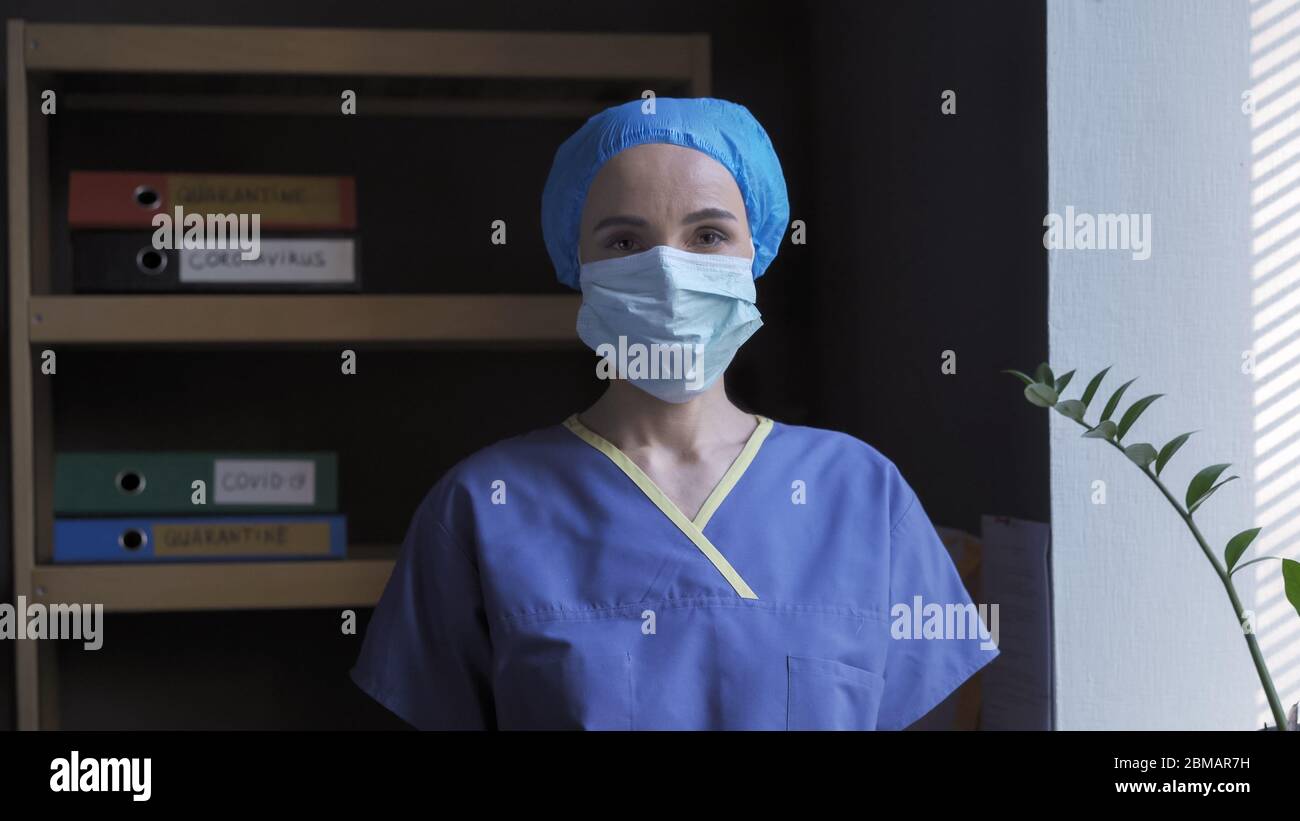 Female Doctor standing in her office. Serious woman in protective mask and blue uniform looks at the camera while standing against a background of Stock Photo